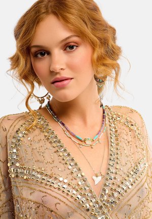 Blouse transparente en sequins dorés avec des perles finement travaillées, superposée de colliers colorés en perles et de boucles d'oreilles ornées au design floral.