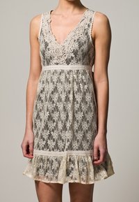 Valkoinen pitsimekko vaaleassa beessä, jossa on V-pääntie, kukkakuvio ja aallotettu helma. Mekossa on muotoiltu yläosa ja liehuva hame.