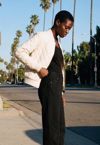 Donna che indossa una giacca trapuntata bianca sopra una maglia nera e pantaloni neri, in piedi su una strada soleggiata fiancheggiata da alte palme.