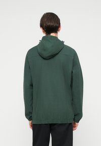 Ciemnozielona kurtka z kapturem, z regulowanym sznurkiem, elastycznymi mankietami i luźnym krojem. Gładka tekstura i minimalistyczny design.