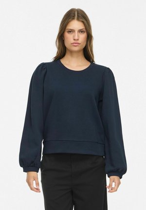 Femme aux longs cheveux bruns porte un haut à manches longues bleu marine avec des épaules bouffantes et un pantalon noir sur un fond blanc uni.