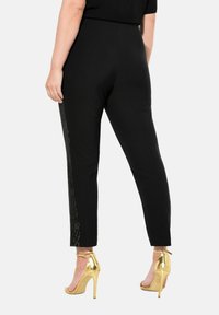 Pantalons noirs à coupe ajustée, avec une bande latérale texturée dorée, associés à des sandales à talons hauts en métal doré.