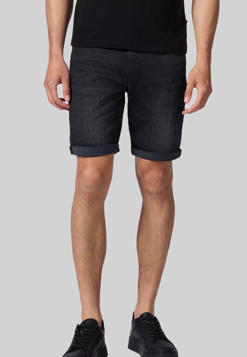 Ciemnoniebieskie szorty dżinsowe z podwiniętymi nogawkami, o prostym kroju i minimalistycznym designie. Noszone z czarnymi trampkami na szarym tle.