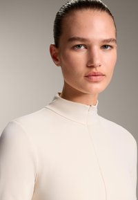 Top beige a collo alto con una texture liscia e dettagli di cucitura sottili, dotato di una chiusura con cerniera corta al collo. Primo piano su un volto.