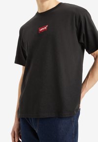 Man som bär en svart Levi's t-shirt med röd logotyp på bröstet och blåjeans, står med en hand i fickan.