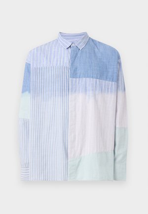 Mees, kes kannab musta nokamütsi, sinise ja valge triibulise patchwork-nööpidega särgi, halli cargo-pükse ja musti tossusid, seisab käega taskus.