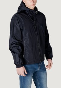 Giacca nera in nylon leggero con zip frontale, polsini elasticizzati e cappuccio. Presenta un piccolo logo sul petto e tasche laterali con zip.