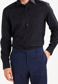 Camicia nera a maniche lunghe con colletto button-down, tasca frontale e texture liscia, abbinata a pantaloni eleganti blu navy.