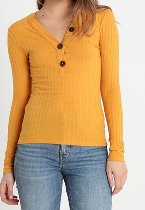 Kvinde iført en sennepsgul ribbet langærmet top med tre mørke knapper og lyseblå denimjeans.