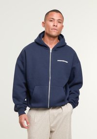 Man wearing a navy blue zip-up hoodie with "PROHIBITED" text and light beige pants, standing with one hand in pocket against plain background.
