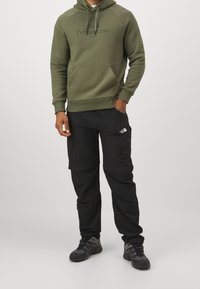 Olijfgroene hoodie met een buidelzak en logo, gecombineerd met zwarte cargobroeken met zijzakken en een logopatch, gedragen met grijze schoenen.