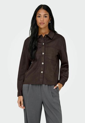 Chemise en suede marron à boutons avec des manches longues, deux poches poitrine et des garnitures argentées, associée à un pantalon gris.