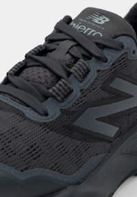 Chaussure de sport noire avec un dessus texturé, œillets renforcés, lacets épais et logo New Balance gris prominent. Dispose d'une semelle rembourrée.