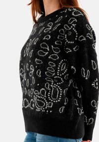 Zwarte gebreide trui met witte abstracte patronen, een relaxte pasvorm, dikke manchetten en een ronde halslijn. Zachte textuur met een fuzzy uitstraling.
