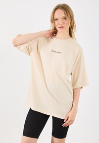 Beige oversized t-shirt van zachte katoen, met korte mouwen en een ronde halslijn. Een kleine zwarte tekst, "Cᴀʀᴇ", is gedrukt op de borst.