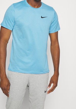 Homme portant un t-shirt Nike bleu clair et un jogging Nike gris, debout devant un fond clair uni.