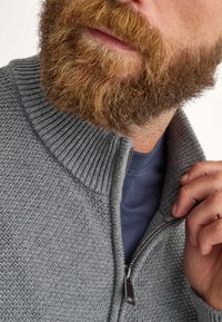 Felpa grigia con zip, caratterizzata da un motivo a maglia testurizzata e colletto a coste. Sotto si intravede una camicia blu navy, con un dettaglio della zip argentato.