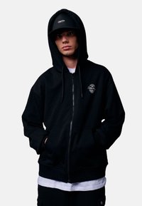 Zwarte vest met ritssluiting van katoenmix, met een voorzak, capuchon met trekkoord en een klein logo op de linker borst. Model draagt een pet.