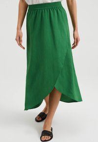 Grüne, strukturierte Maxiröhre mit elastischem Taillenbund und Wickeldesign. Tragen mit schwarzen, gewebten Rutsch-Sandalen.