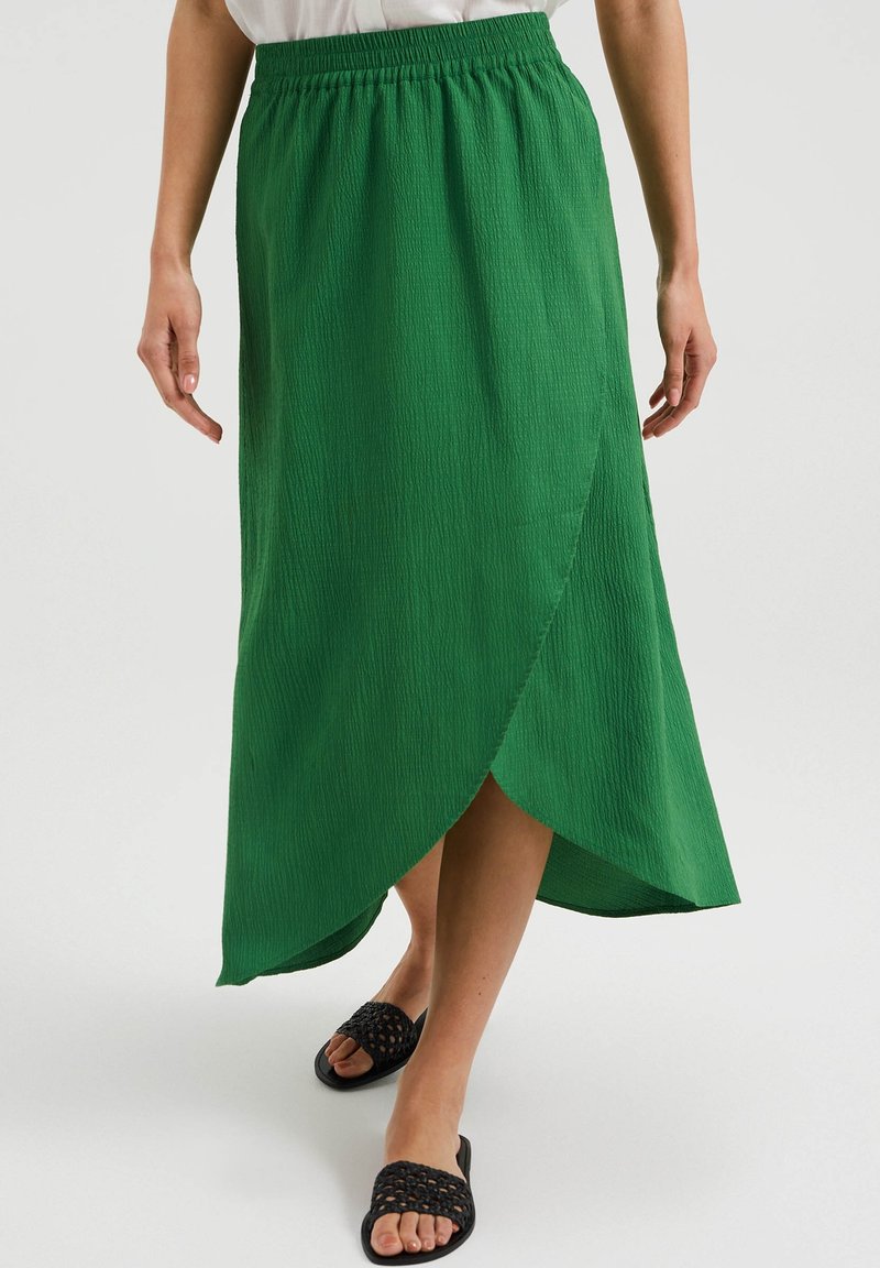 Grüne, strukturierte Maxiröhre mit elastischem Taillenbund und Wickeldesign. Tragen mit schwarzen, gewebten Rutsch-Sandalen.