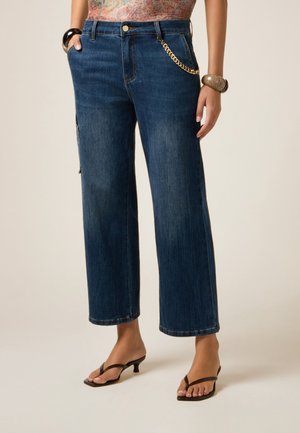 Femme portant un jean large bleu foncé avec un détail chaîne dorée, un haut fleuri, des sandales à talons marron et des bracelets épais.