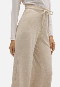 Pantalones de pierna ancha de color beige, hechos de una tela suave, con una cinturilla alta, cierre con cordón y una sutil textura, además de bolsillos laterales.