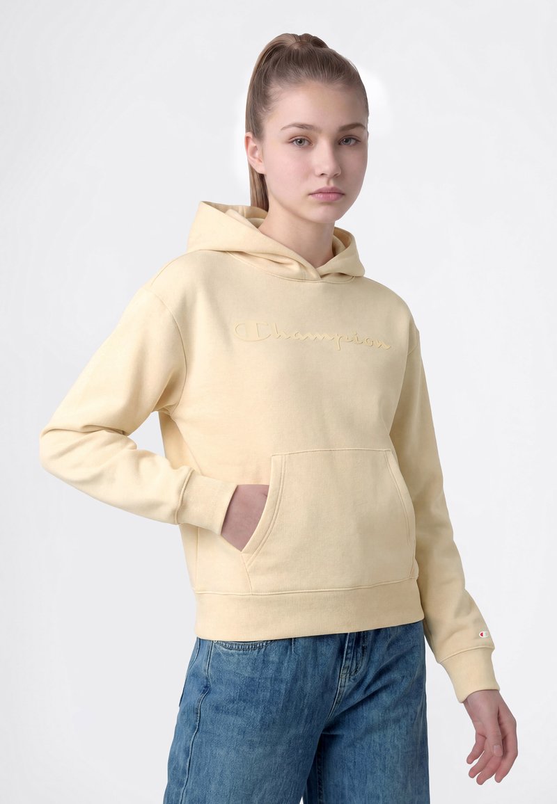 Jeune femme avec une queue de cheval porte un sweat à capuche beige Champion et un jean bleu, debout avec une main dans la poche du sweat à capuche sur un fond uni.