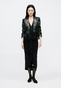 Črn usnjen blazer z izrezom iz čipke, kombiniran s črno nabrano obleko. Model nosi črne nogavice in koničaste balerinke. Preprost, eleganten dizajn.