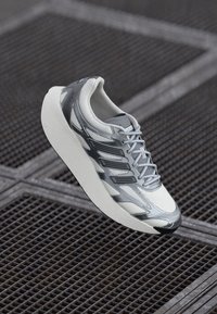 Hvid og grå sportsko med tyk sål og mønstret mesh-overdel, svævende over en metalristsurface.