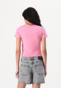 Kvinna med långt lockigt hår som bär en kortärmad rosa topp och höga grå denimshorts med bakfickor, stående mot vit bakgrund.