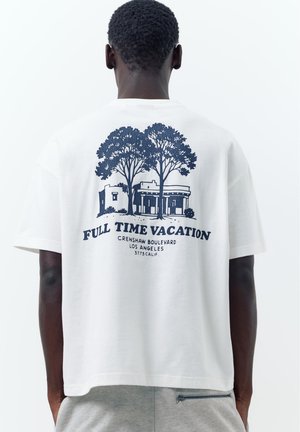Uomo che indossa una t-shirt bianca con grafica blu navy di alberi, edifici e testo "Full Time Vacation Crenshaw Boulevard Los Angeles."