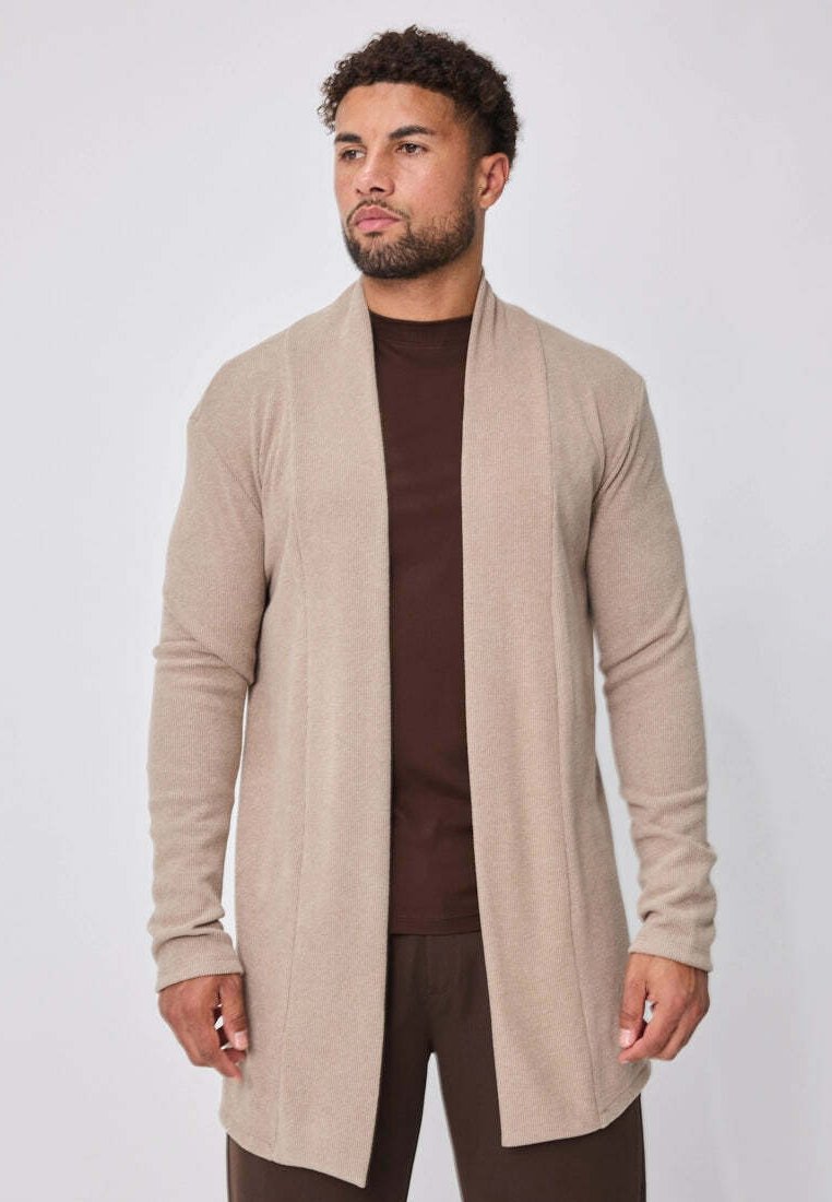 Homme portant un cardigan beige ouvert sur un t-shirt et un pantalon marron foncé, debout devant un fond uni gris clair.