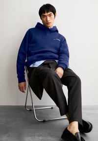 Jonge man die op een metalen stoel zit, gekleed in een blauwe hoodie, een gestreept shirt, donkere broeken en zwarte instappers, tegen een effen muur.