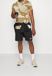 Camiseta de Nike en color beige con patrones abstractos, pantalones cortos negros y zapatos beige. Sosteniendo una botella de agua, con una chaqueta negra atada alrededor de la cintura.