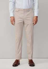 Light beige tailored trousers made of textured fabric, featuring a side pocket, flat front, and rolled cuffs, paired with dark brown shoes.
