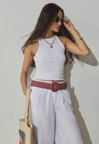 Top blanco sin mangas, cinturón trenzado burdeos, pantalones blancos holgados, collar de cadena dorada, gafas de sol redondas, bolso beige con detalle colorido.