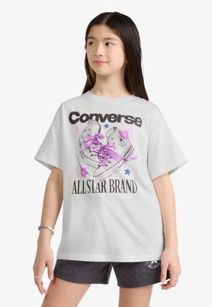 Mladé dievča nosí biele oversized tričko s grafikou tenisky Converse a čierne šortky, pozerá sa do strany.