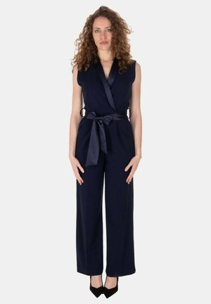 Donna con capelli ricci che indossa una tuta blu navy senza maniche con una cintura di raso annodata in vita e tacchi neri a punta, in piedi ferma.