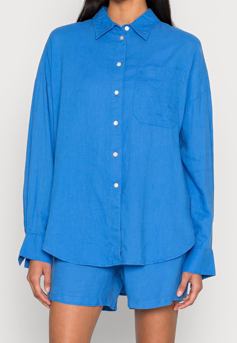 Femme portant une chemise bleue à manches longues et amples, boutonnée, avec une poche poitrine et un short bleu assorti sur un fond uni.