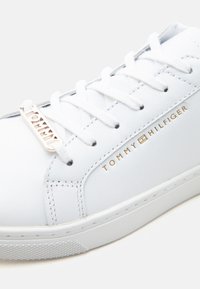 Άσπρο δερμάτινο αθλητικό παπούτσι με λευκά κορδόνια, χρυσό λογότυπο "Tommy Hilfiger" στο πλάι και χρυσή ετικέτα κορδονιών με το εμπορικό σήμα σε λευκή σόλα.
