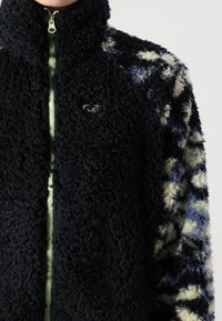 Fleece bunda s vysokým límcem, černým strukturovaným tělem, rukávy s vícebarevným vzorem a světle zeleným zipem s malým logem.