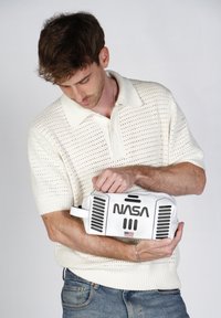 Bolsa blanca con el logo de "NASA"; presenta detalles en negro y ranuras de ventilación; sostenida por una persona que lleva una camiseta polo de punto ligero y texturizado.