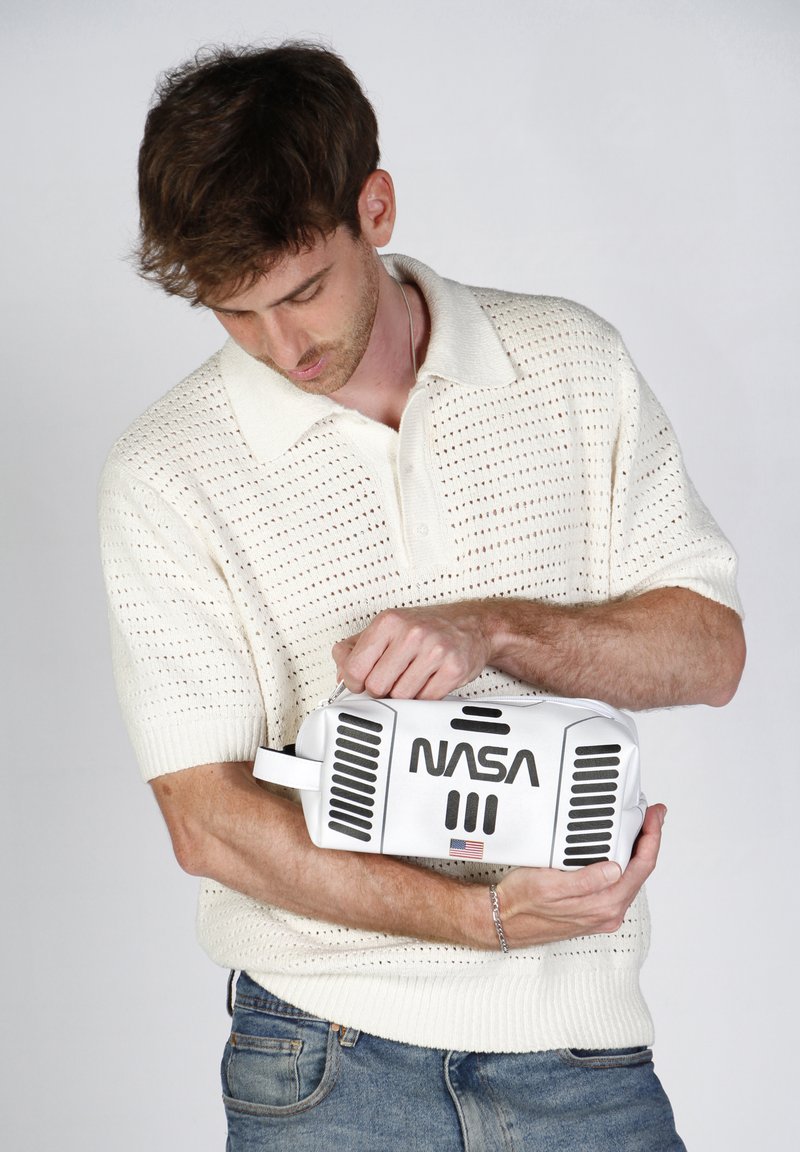 Bolsa blanca con el logo de "NASA"; presenta detalles en negro y ranuras de ventilación; sostenida por una persona que lleva una camiseta polo de punto ligero y texturizado.