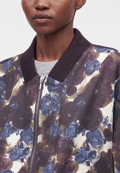 Gros plan d'une personne portant une veste bomber à motif floral avec col en côtes violet et fermeture éclair sur une chemise gris clair.