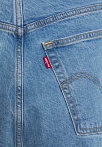 Jeans de mezclilla azul claro con costuras naranjas contrastantes y una etiqueta roja en el bolsillo trasero. La textura es suave con un ligero desgaste.