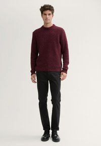 Burgunderfarbener Strickpullover mit strukturiertem Muster, rundem Ausschnitt und gerippten Bündchen, kombiniert mit schwarzen Hosen und schwarzen Schuhen.