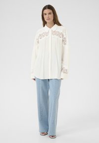 Elfenbeinfarbige Bluse mit langen Ärmeln, bestickten Spitzelementen, Kragen und Knopfverschluss, kombiniert mit hellblauen weit geschnittenen Jeans. Glatte Textur, lockere Passform.