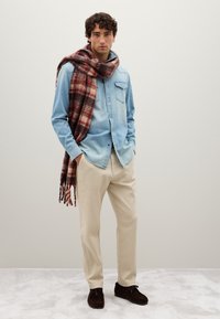 Man in een lichtblauw denim shirt, beige broek, donkerbruine schoenen en een grote rood en bruine geruite sjaal over zijn schouder gedrapeerd.
