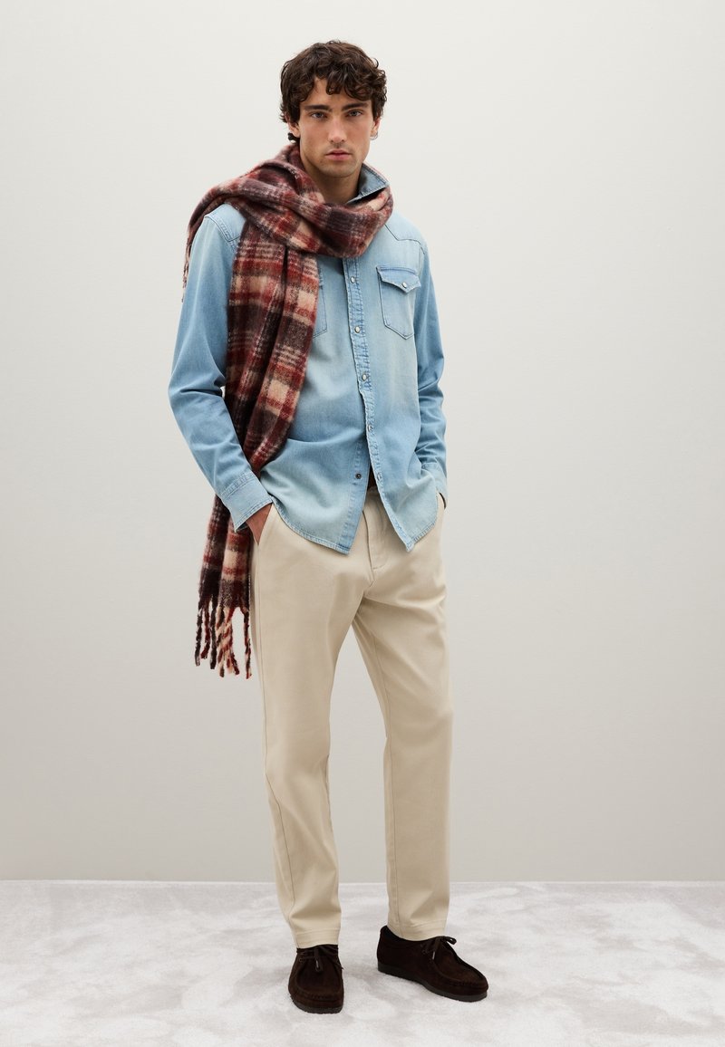 Man in een lichtblauw denim shirt, beige broek, donkerbruine schoenen en een grote rood en bruine geruite sjaal over zijn schouder gedrapeerd.