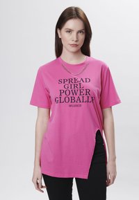 Camiseta oversized rosa con mangas cortas, abertura lateral y texto impreso en negro que dice "SPREAD GIRL POWER GLOBALLY INFLUENCER."